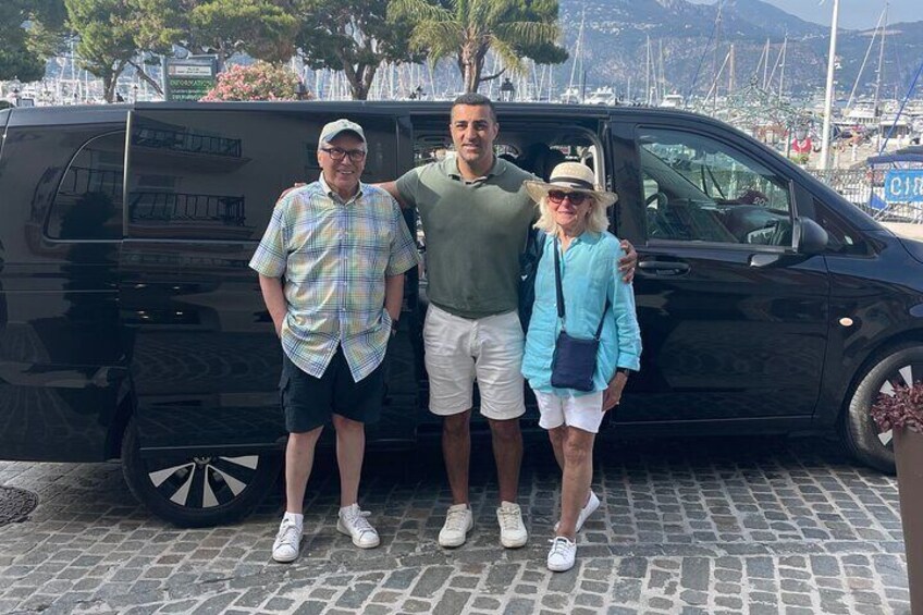 Three people standing in front of a black van on a cobblestone street with a marina and boats in the background. 1