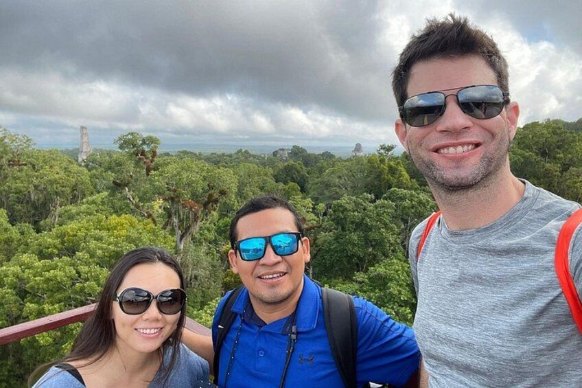 Three people standing on a wooden platform with a forested landscape in the background. 1