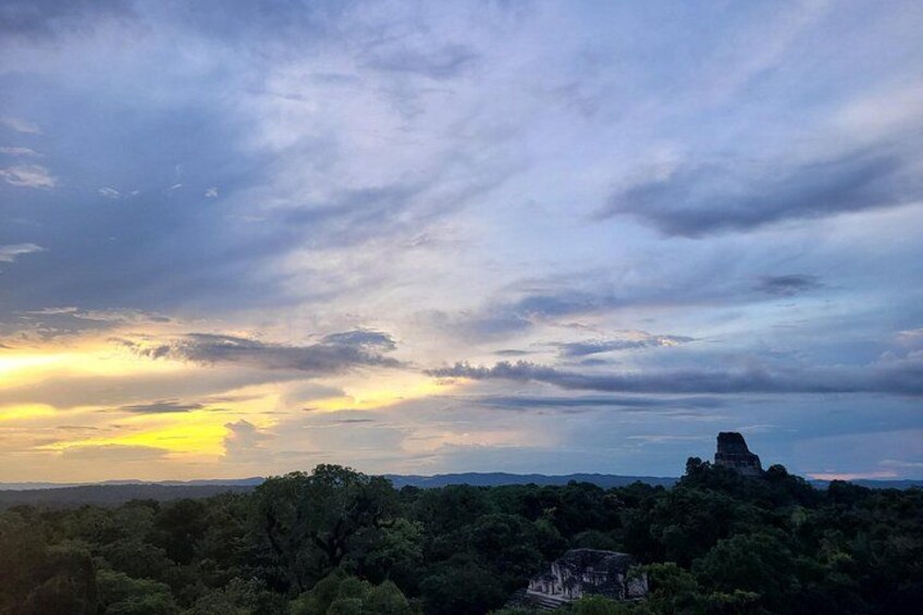A sunset over a forested landscape with a prominent hill and a small building. 3