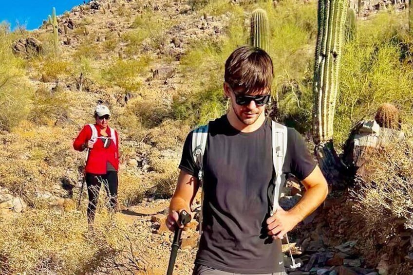 Two hikers on a rocky trail with cacti and sparse vegetation. 3