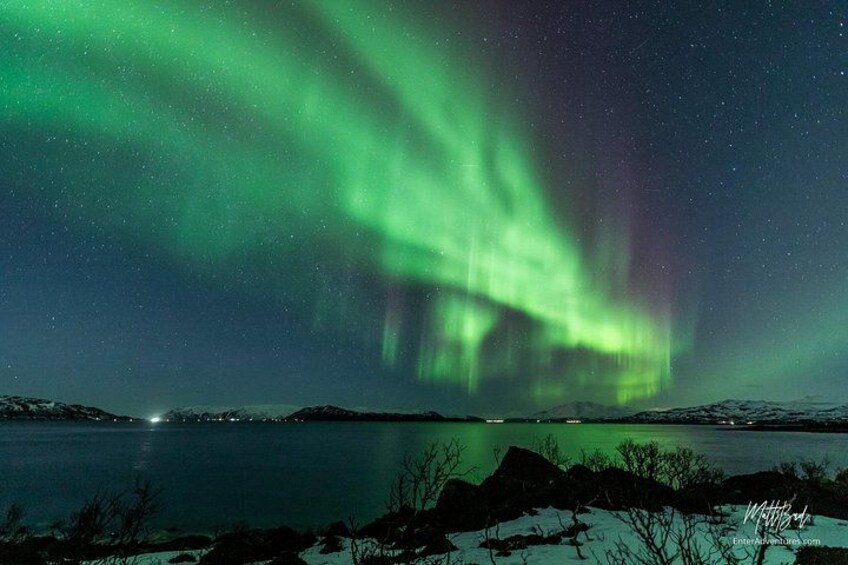 Norrsken över en stilla sjö, med snötäckta klippor i förgrunden. 2