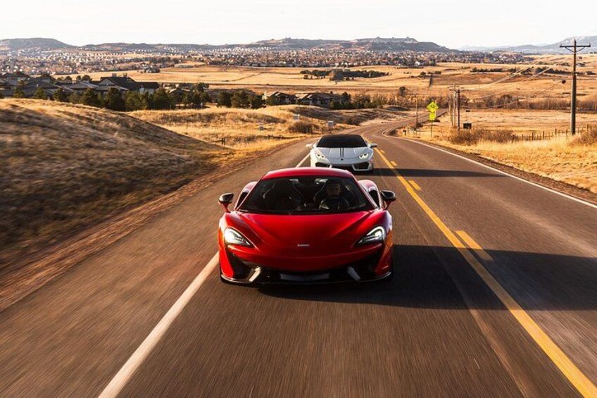 A red sports car driving on a two-lane road with a white car ahead, surrounded by open fields and distant hills. 5