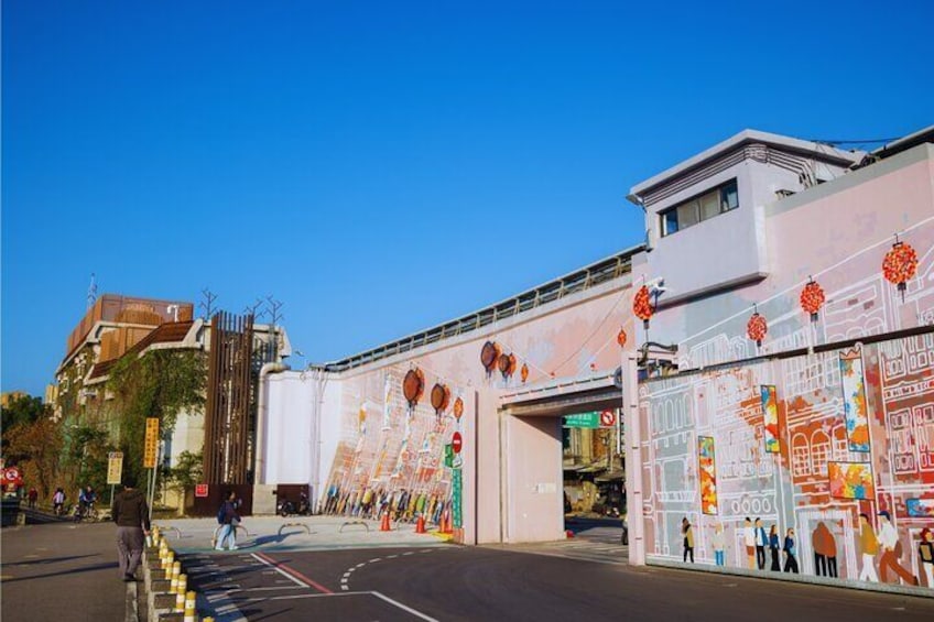 Una calle con un gran mural en un edificio, cielo azul despejado y un paso de peatones. 2