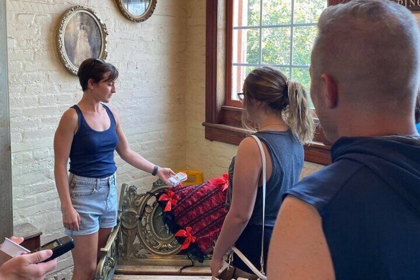 Une femme, vêtue d’un débardeur bleu et d’un short en jean, tient un téléphone et se tient près d’une fenêtre. Une couverture rouge est posée sur une chaise. 1
