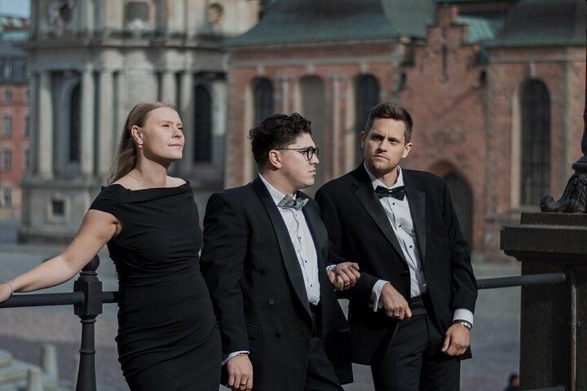 Three individuals in formal attire standing on a balcony with a historical building in the background. 1
