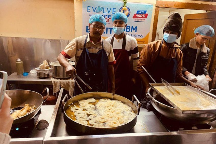 Une cuisine où des chefs préparent des plats, une grande marmite remplie de nourriture et une banderole en arrière-plan. 5