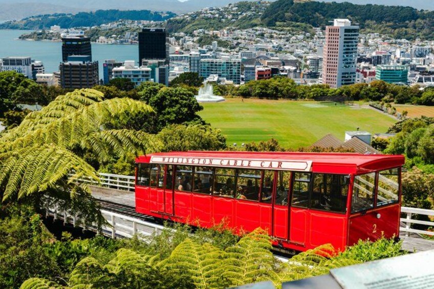 A red cable car with 'BIRMINGHAM CABLECAR' written on it, traveling over a bridge with greenery and a cityscape in the background. 1