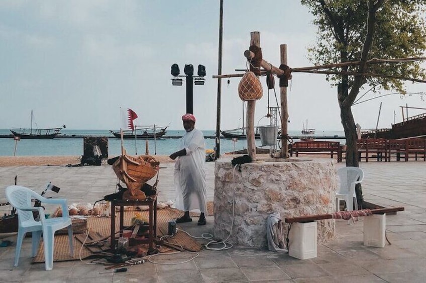 A man in traditional attire standing near a wooden structure with a flag, a beach, and boats in the background. 5