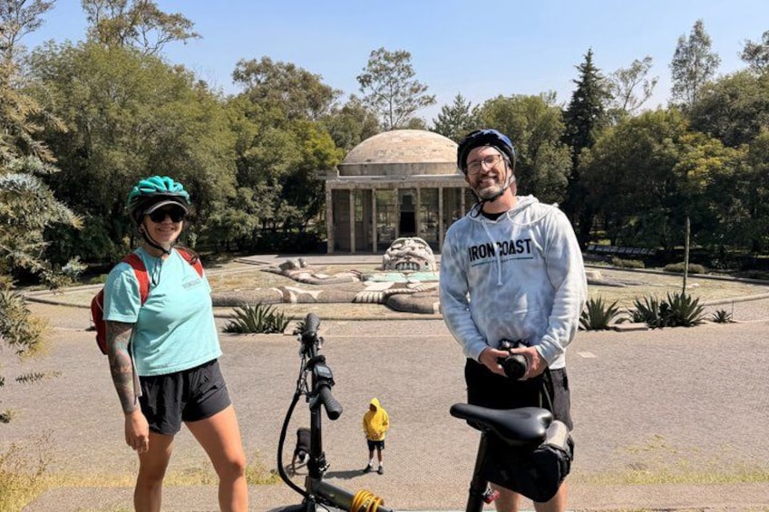 Two people standing in a park with a fountain and a building in the background. 4