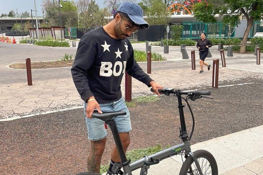 A person adjusting a bicycle on a paved path with bollards, a running person in the background, and a building with a green roof. 5