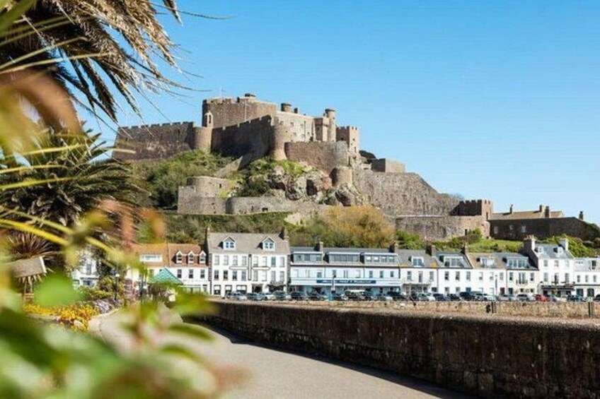 A coastal town with a historic castle on a hill, a promenade with buildings, and palm trees. 4