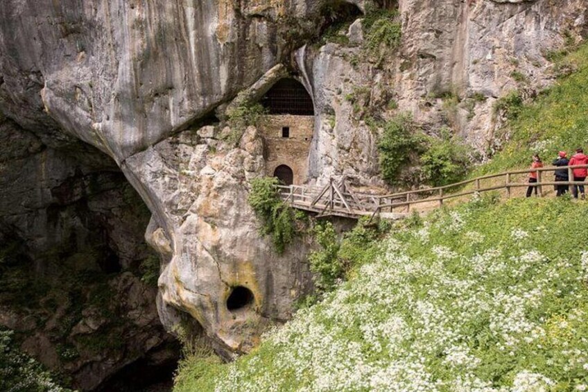 A cave entrance with a wooden bridge, a small building, and hikers on a trail. 2