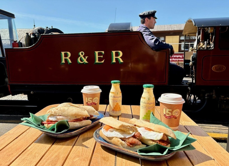 A vintage train with 'R&ER' on the side, a dining table with sandwiches and drinks, and a person in uniform. 1