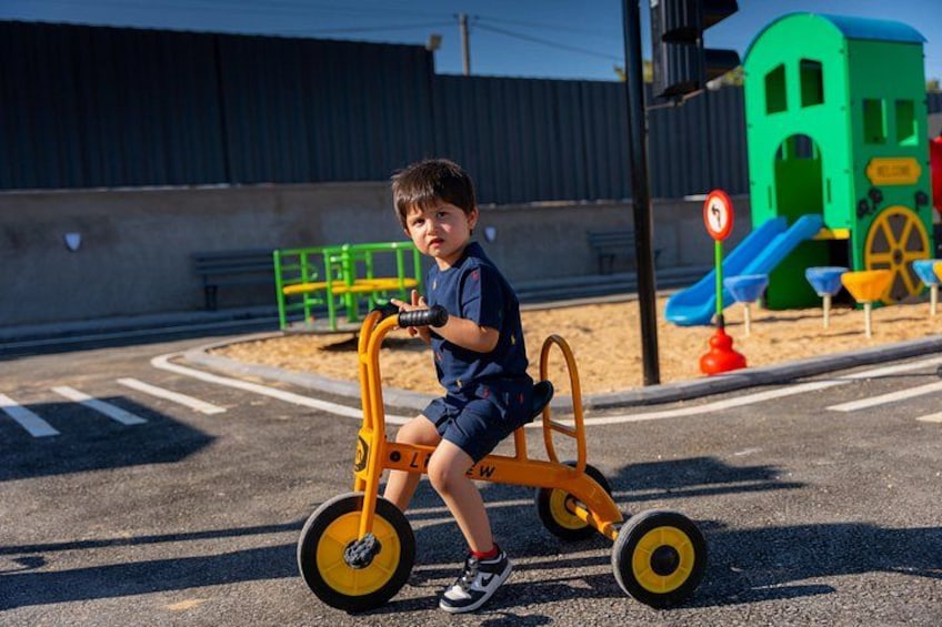 A child riding a tricycle on a street with a playground in the background. 1