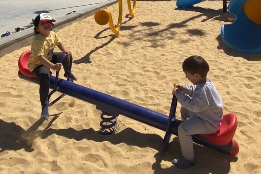 Deux enfants jouent sur des balançoires à bascule sur une plage de sable, avec des jeux pour enfants en arrière-plan. 3