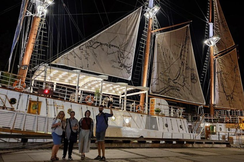 Ett stort segelfartyg låg förtöjt vid en hamn, och folk stod på däck. 2
