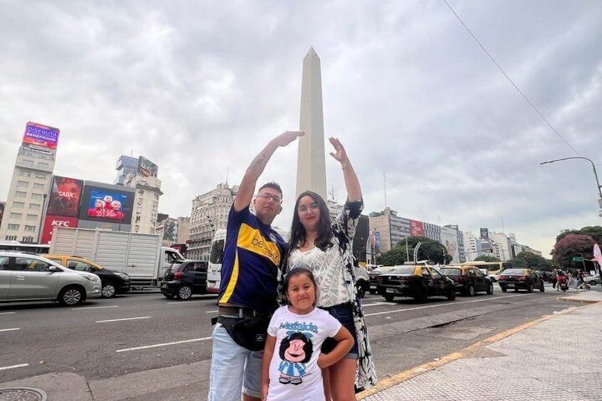 Una familia posando junto a un monumento alto en un entorno urbano, con edificios y vehículos al fondo. 3