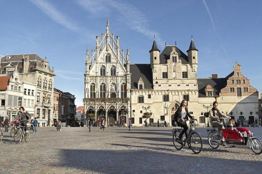 Un edificio histórico con arquitectura elaborada, una plaza adoquinada y gente andando en bicicleta. 3