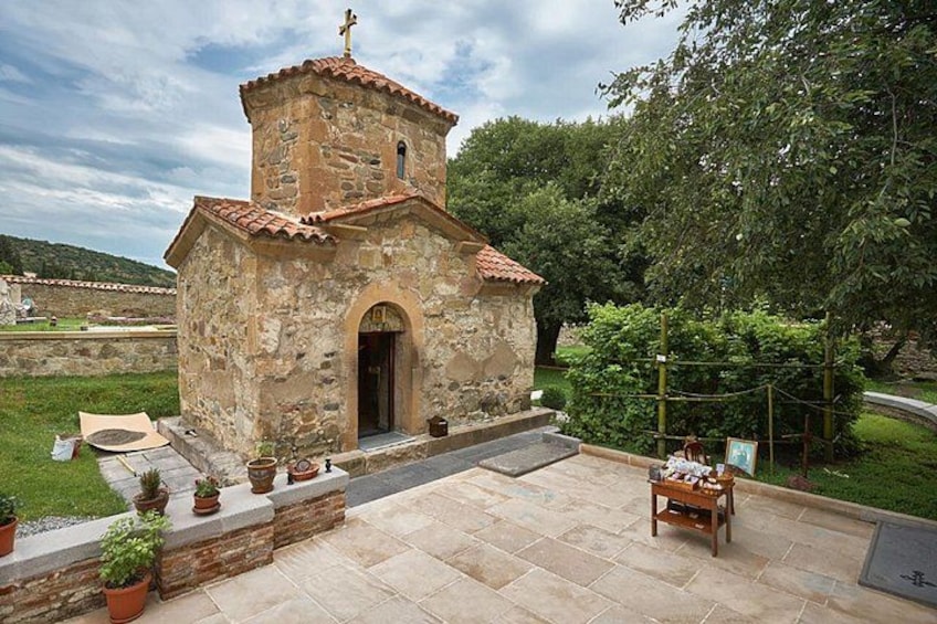 A small stone church with a bell tower, surrounded by a paved courtyard with potted plants and a wooden bench. 3