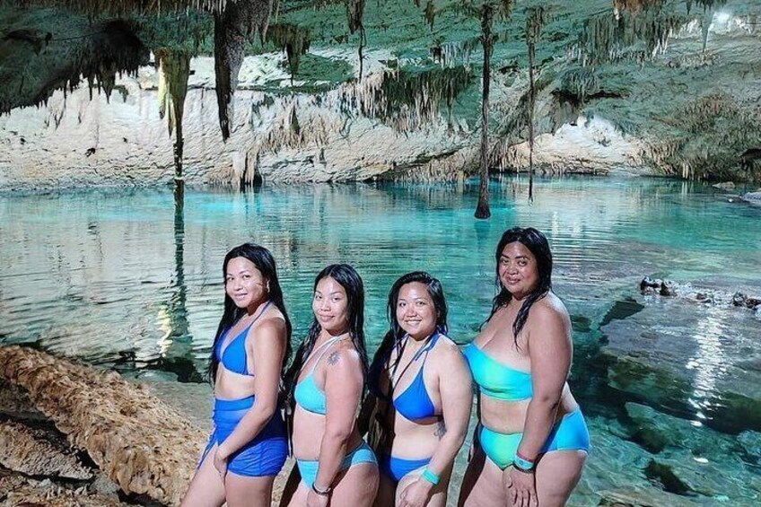 Un grupo de mujeres con trajes de baño azules de pie junto a un cuerpo de agua azul y cristalina, rodeado de terreno rocoso y árboles. 2