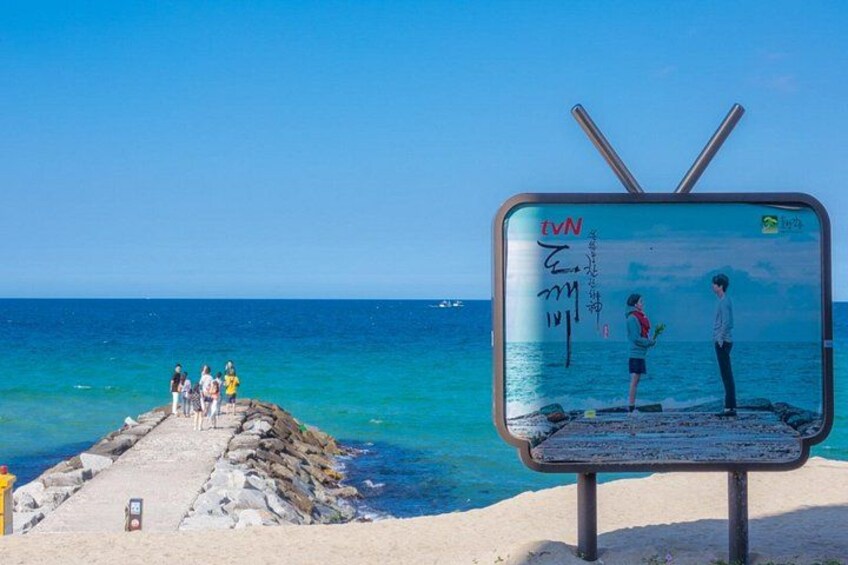 Een strand met een televisiescherm waarop een beeld te zien is, een rotsachtig pad en de oceaan. 2