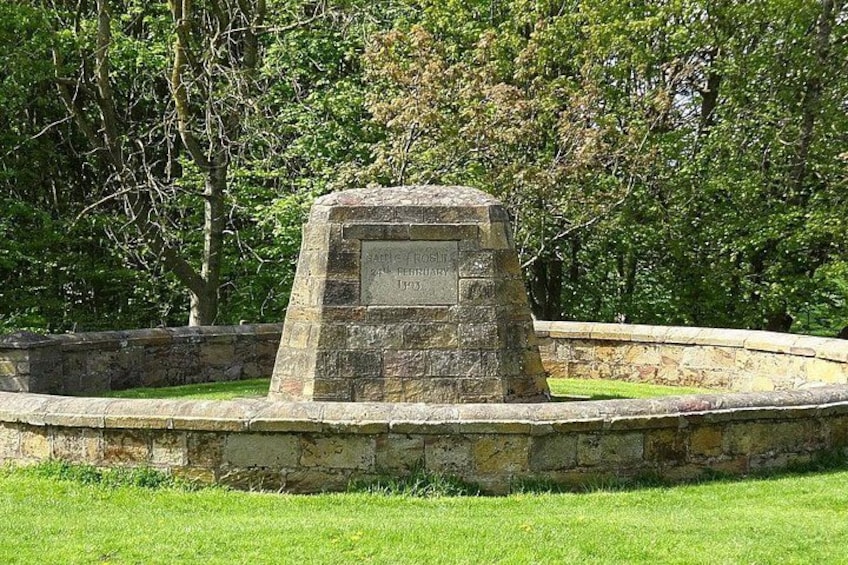 A stone monument with an inscription, surrounded by a circular stone wall, set in a grassy area with trees in the background. 2
