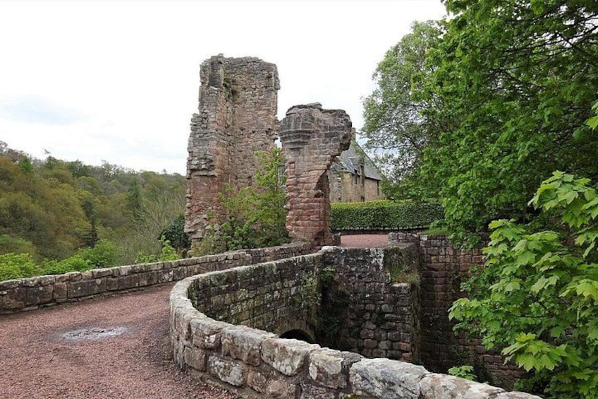 A historic stone bridge with a circular stone wall, leading to a ruined castle structure amidst a forested landscape. 3