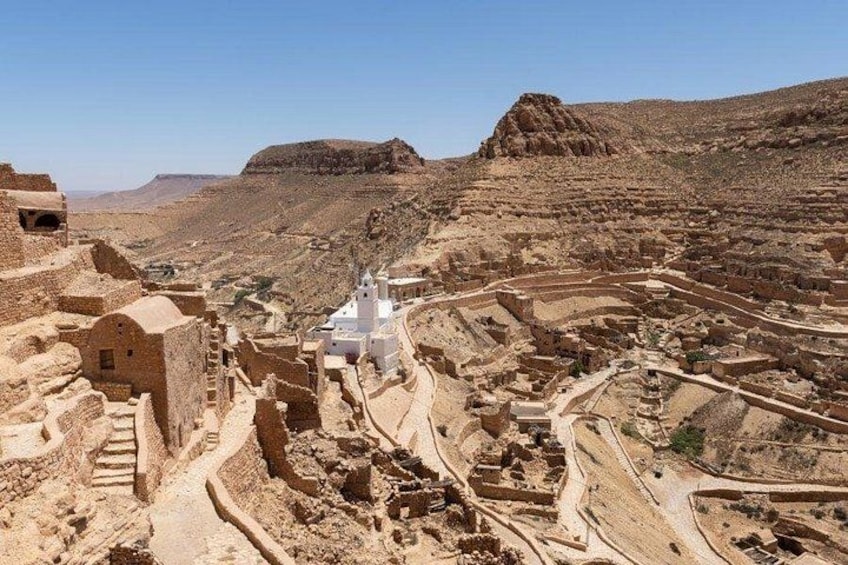 An ancient archaeological site with ruins, including a white building with a dome and a large open area with stone structures. 2
