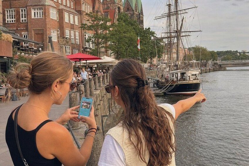 Dos personas fotografiando un barco de vela alto amarrado en el muelle, con edificios al fondo. 5