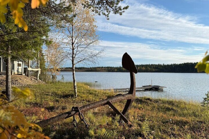 Escena junto a un lago con una escultura rústica, un muelle de madera y una casa. 3