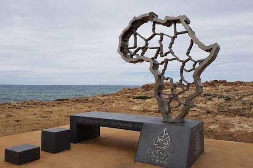 En skulptur i metall som liknar en sprucken jord eller ett träd, placerad på en betongplattform med bänkar, nära en strand med en stenig kustlinje. 1
