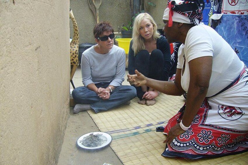 Three people sitting on a mat in a courtyard with a giraffe in the background. 1
