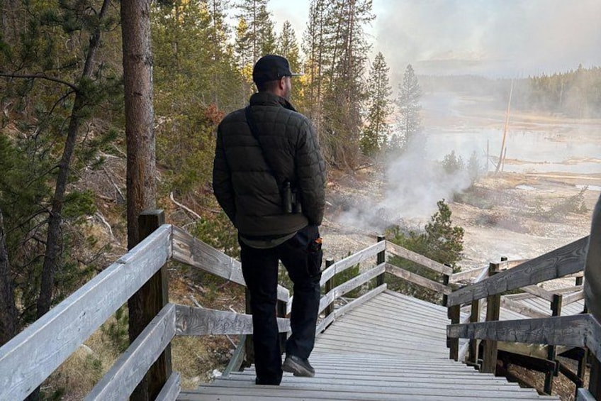 A person standing on a wooden boardwalk overlooking a geothermal area with steam rising from the ground. 2