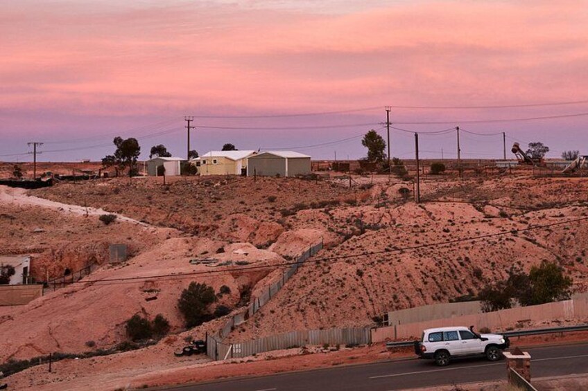 A desert landscape with sparse vegetation, a dirt road, a white car, and a few buildings. 3