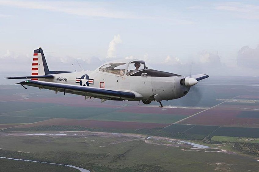 A vintage military aircraft in flight over a landscape with fields and a river. 3