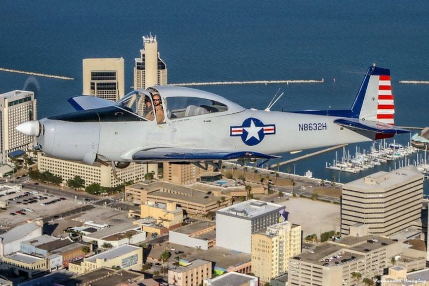 A vintage military aircraft flying over a cityscape with buildings and a marina. 2