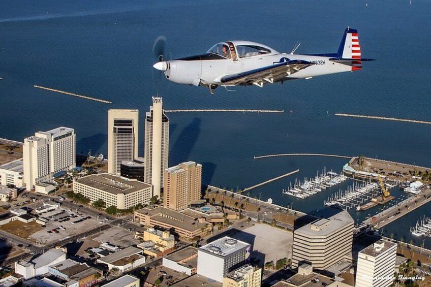 A small aircraft flying over a coastal city with high-rise buildings and a marina. 1