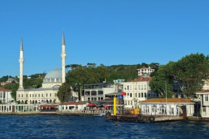 A waterfront view of a historic building with two minarets, a mosque, and a modern building with a yellow sign. 2