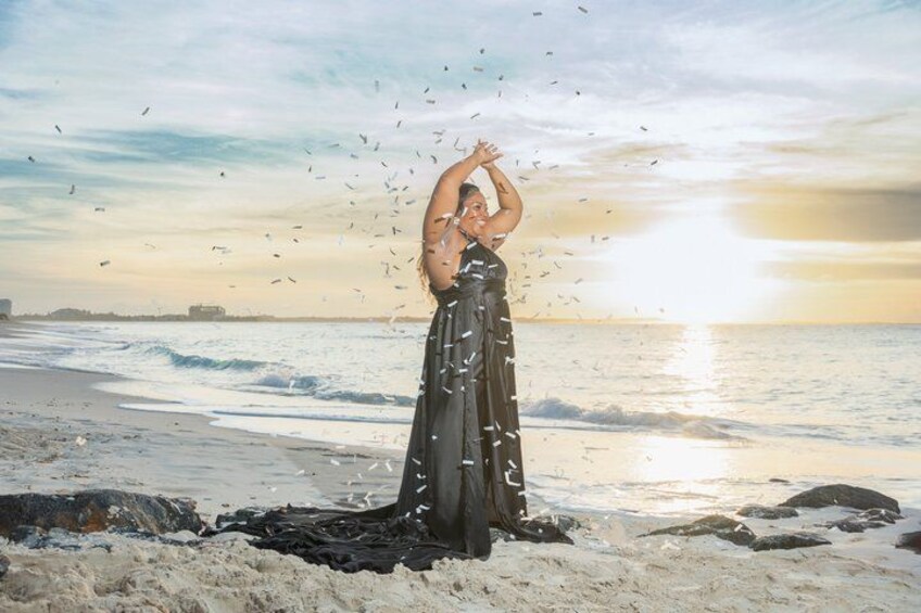 A person in a long black dress standing on a beach with waves and a sunset in the background. 4