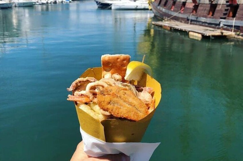 A hand holding a food container with fried items and a lemon wedge, with a body of water and boats in the background. 1