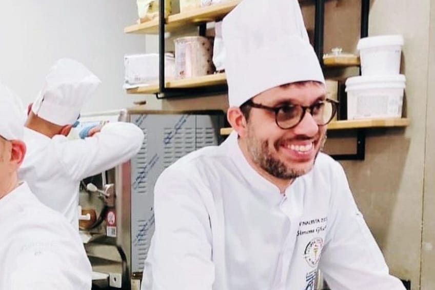 Two chefs in a kitchen, one adjusting his hat, with shelves of kitchenware in the background. 2