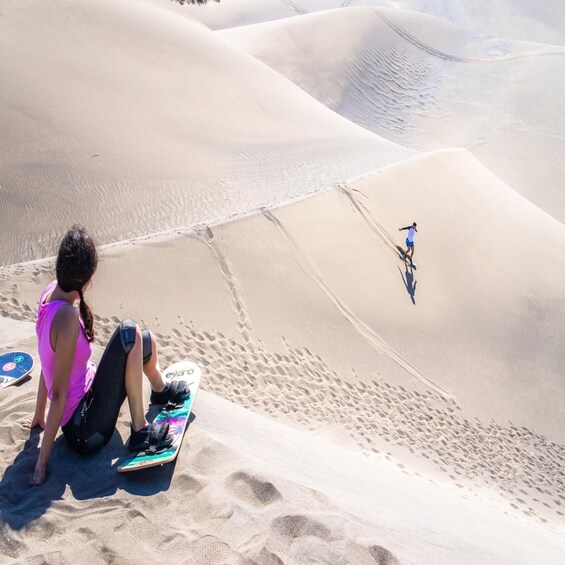 A person sitting on a sand dune with a skateboard, another person riding a skateboard down the dune. 1