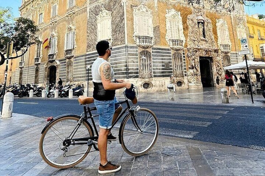 Una persona de pie junto a una bicicleta frente a un edificio histórico con arquitectura ornamentada. 2