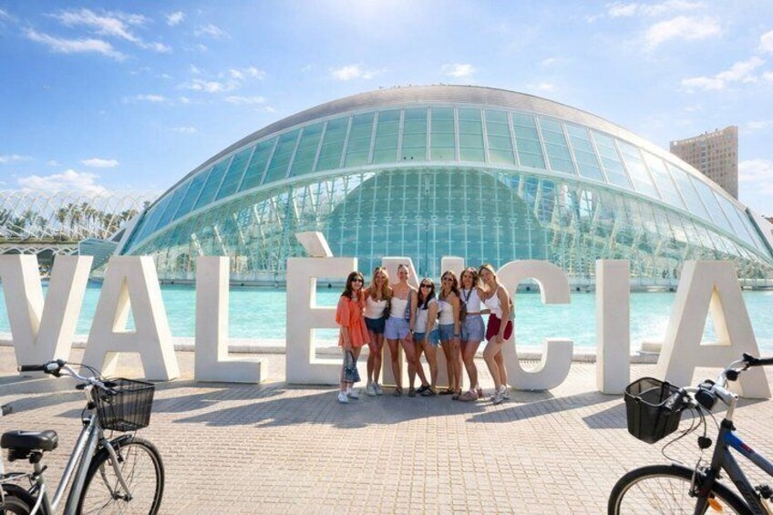 Eine Gruppe von Personen steht vor einem großen Schild mit dem Wort "VALENCIA" und einem modernen Glasgebäude im Hintergrund. 1