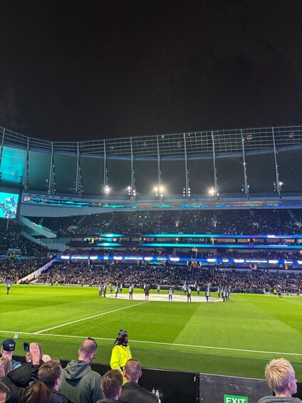 Un estadio con múltiples niveles de asientos, un campo verde y una pantalla grande mostrando un evento. 3