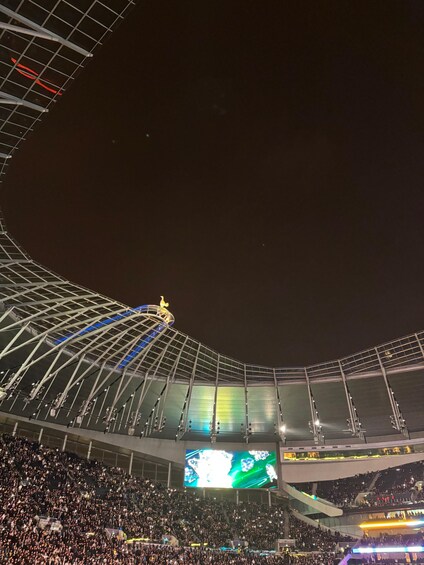 Un estadio con una gran multitud, una estructura de techo curva y una pantalla gigante mostrando una imagen. 2