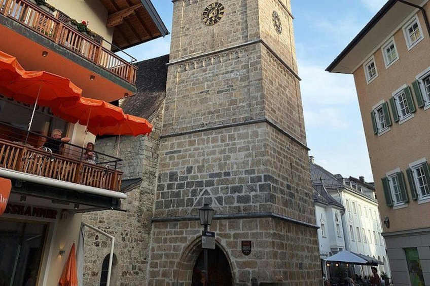 Un'antica torre in pietra con una torre dell'orologio, un balcone con ombrelloni arancioni e edifici adiacenti. 1