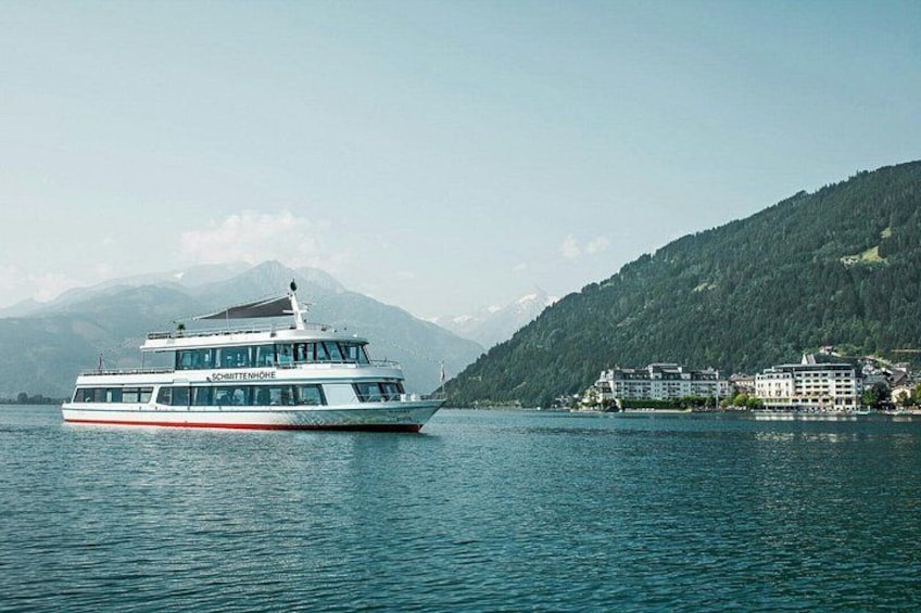 Una nave da crociera bianca con decorazioni rosse che naviga su acque calme, con montagne sullo sfondo e una piccola cittadina con edifici sulla costa. 5