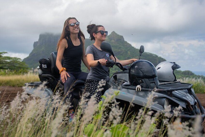 Two people on a motorbike in a grassy area with a mountain in the background. 3