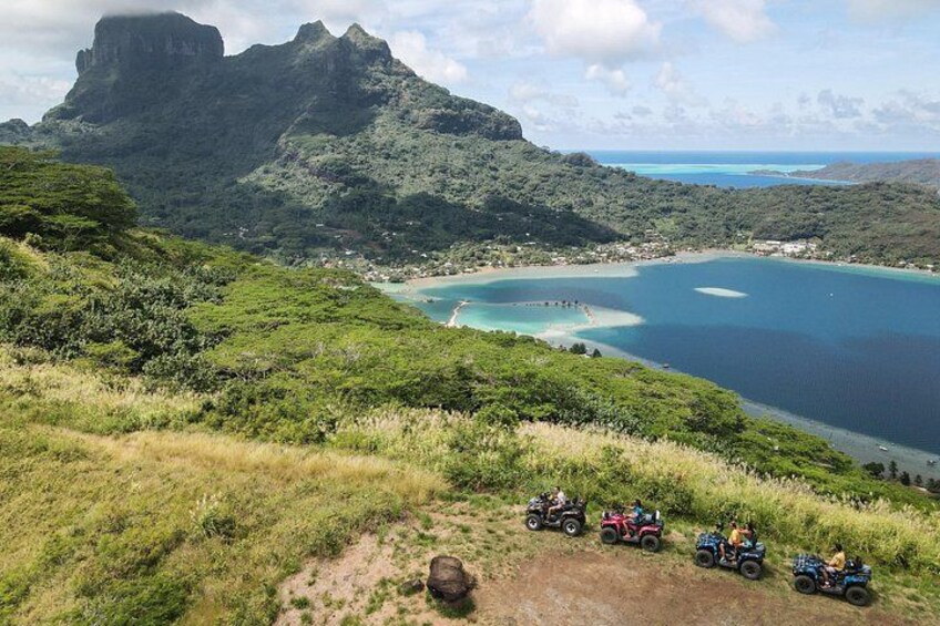 A coastal landscape with a large mountain, a beach, and four off-road vehicles on a dirt path. 1